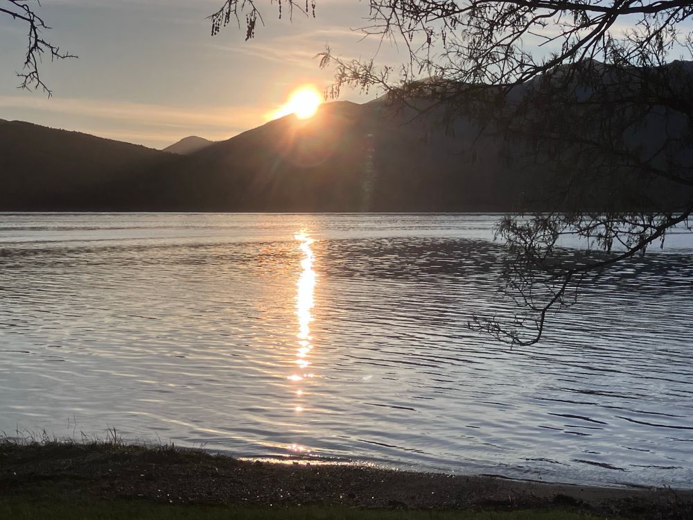 The Sun sets behind mountains in front of which it is reflected in the waters of a lake