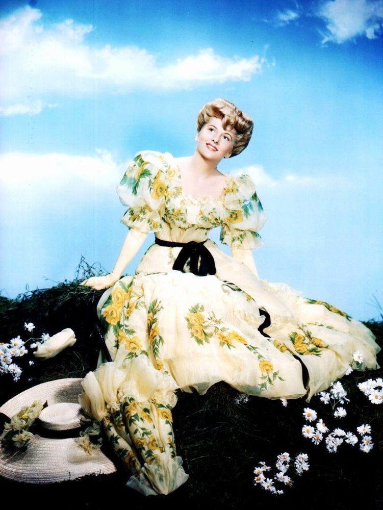 Joan Fontaine in a color promo photo for her 1948 musical with Bing Crosby, "The Emperor Waltz". She wears an Edwardian-style flowered gown with puffy short sleeves and a black sash around her waist, with long white gloves; a wide-brimmed, flowered hat and parasol with the same flowered design as her dress rest at her feet, and her blonde hair is done up in a "Gibson Girl" pompadour.