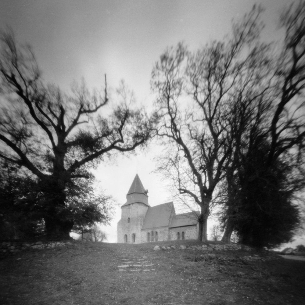 Schwarzweissfoto, etwas unscharf und kontrastarm, superweitwinklig. Bäume mit wenig oder ganz ohne Laub rahmen eine alte Dorfkirche, die auf einem kleinen Hügel steht, ein.