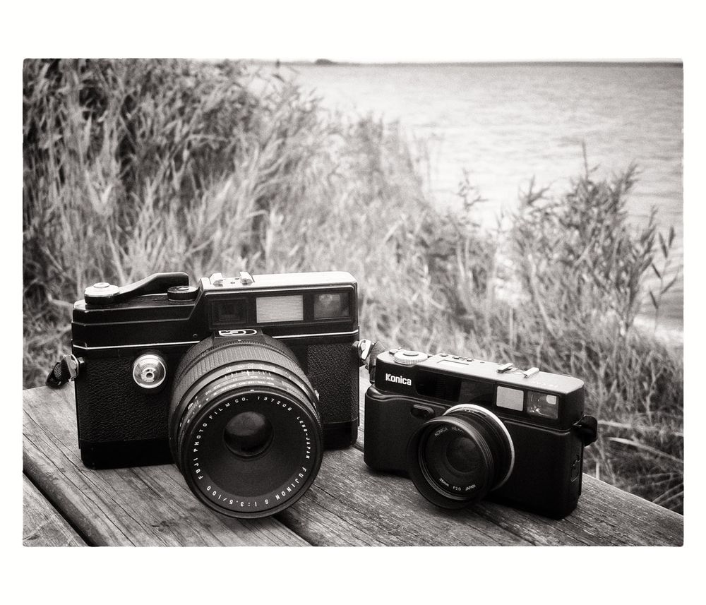 Schwarzweißfoto, zwei Kameras stehen nebeneinander auf einem Holztisch im Freien. Die linke Kamera ist offensichtlich älter und deutlich größer als die Sucherkamera rechts.