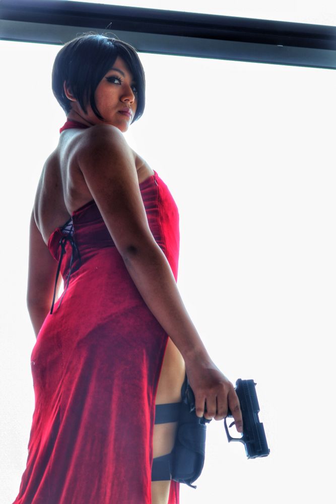 A person cosplaying as Ada Wong from Resident Evil, seen from the back. They have a black bob haircut and are looking over their left shoulder at the camera. They're wearing a red, high-slit dress with a lace-up back and a black leg holster with a handgun. The person is holding a handgun in their right hand, aimed downward. The background is a bright, white light.