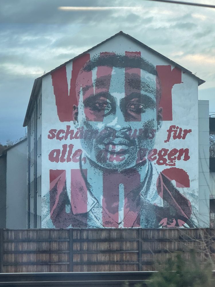 zu sehen ist eine Hausfassade im Frankfurter Stadtteil Niederrad. Auf der Fassade ist ein großes Bild gemalt. Zu sehen ist das Porträt von Tony Yeboah mit der Inschrift „Wir schämen uns für alle gegen uns!“