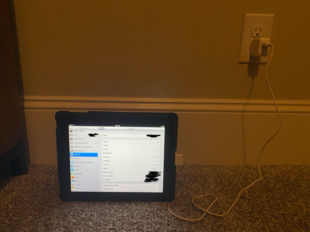 A 2010 first generation Apple iPad, showing its About page. The device has an almost drained battery, and is running off the official 10 W adapter and cable.
