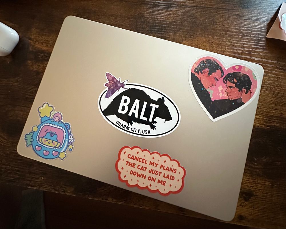 Shot of my laptop and it’s growing sticker collection featuring a purple death head moth, a cartoon rendering of Hannibal and will almost missing, a tamagotchi sticker and a sticker in the shape of a cloud that says “cancel my plans the cat just laid down on me”