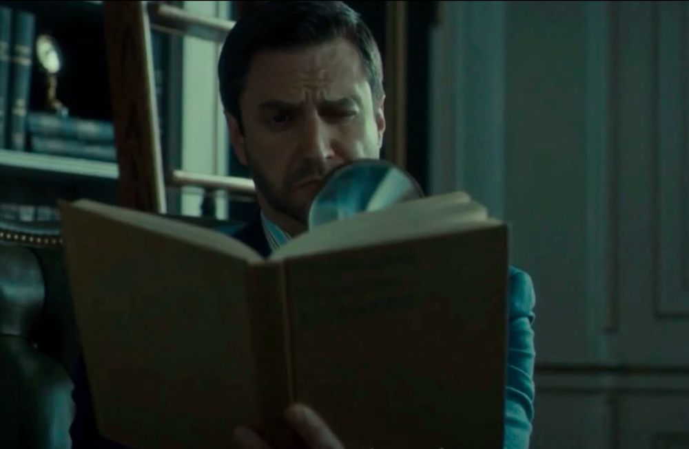 Frederick Chilton sitting at his desk reading a book with a magnifying glass 