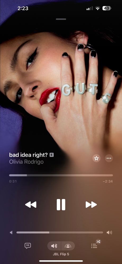 Screenshot of Apple Music olivia Rodrigo’s bad idea right? The album cover is a close up of her with dark hair and a red lip biting her thumb. Her rings spell out GUTS