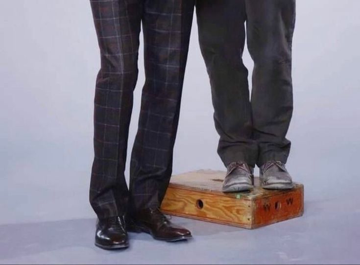 A shot of two men from the thighs down. On the left the man is wearing a plaid suit and wingtips, the man on the right is wearing grey pants, and some scuffed up derby type shoes. He is standing on a box 
