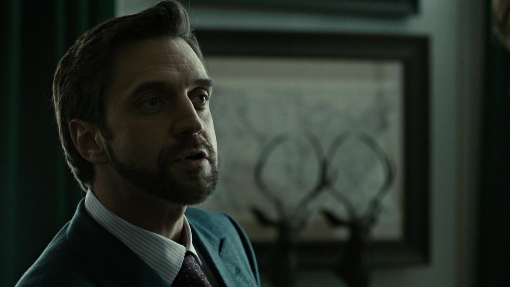 S1 E6 of Hannibal. On the left side in the foreground of the shot a bearded and coiffed Chilton speaks. In the right background two stag bust silhouettes sit on a table behind him 