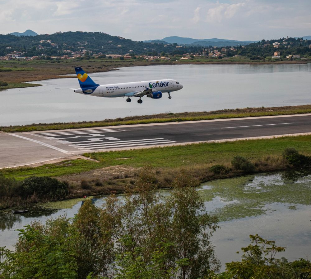 Zeigt ein Flugzeug von Condor im Landeanflug auf die Landebahn von Korfu.
Das besondere ist hier, dass die Lanmdebahn im Wasser liegt, also rechts und links ist Wasser, ganz hinten sieht man ein paar grüne Berge.