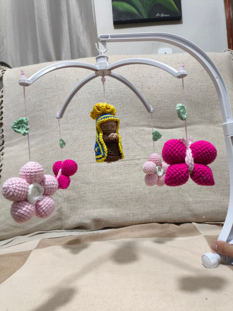 Mobile para bebê com Nossa Senhora no centro. Ao redor, intercaladas, duas flores de 5 pétalas rosas e miolo branco e duas borboletas pink. Todos bonecos de crochê, amigurumis. 
