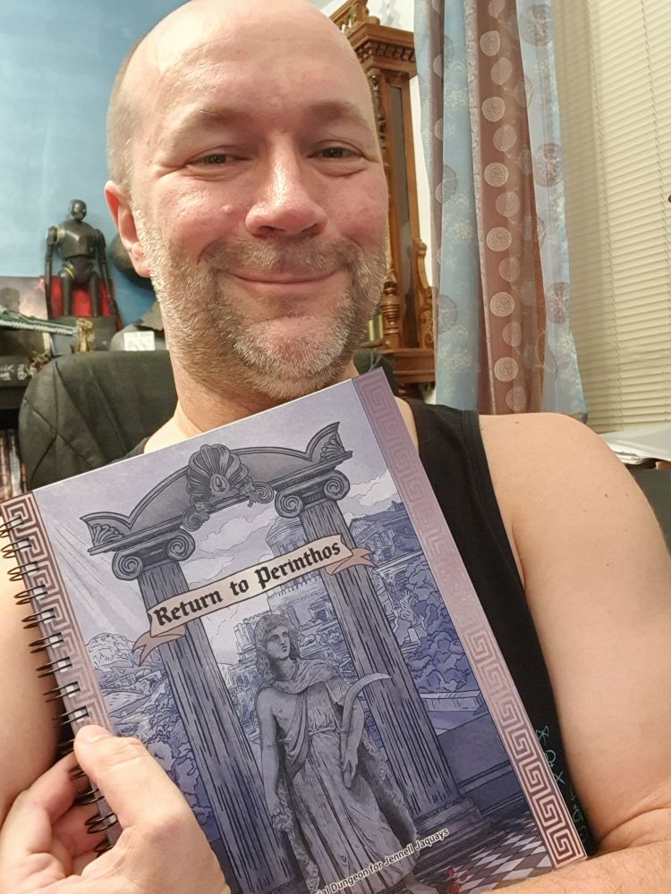 White male (me) smiling into camera while holding up a ttrpg book called Return to Perinthos - A Memorial Dungeon for Jennel Jaquays