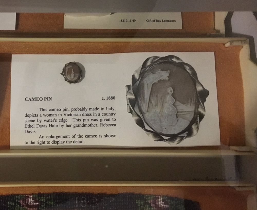 Museum display featuring a came pin in a silver setting on the left with a photo enlargement of the pin on the right. The label reads:
Cameo pin, c. 1880. This cameo pin, probably made in Italy, depicts a woman in Victorian dress in a country scene by water's edge. This pin was given to Ethel Davis Hale by her grandmother, Rebecca Davis. An enlargement of the cameo is shown to the right to display the detail.

Additional notes: The woman figure's eye is carved as a huge dark void disproportionately large for her facial profile. May cause one to speculate as to the specific reason it ended up in a museum rather than continuing its legacy as a family heirloom.