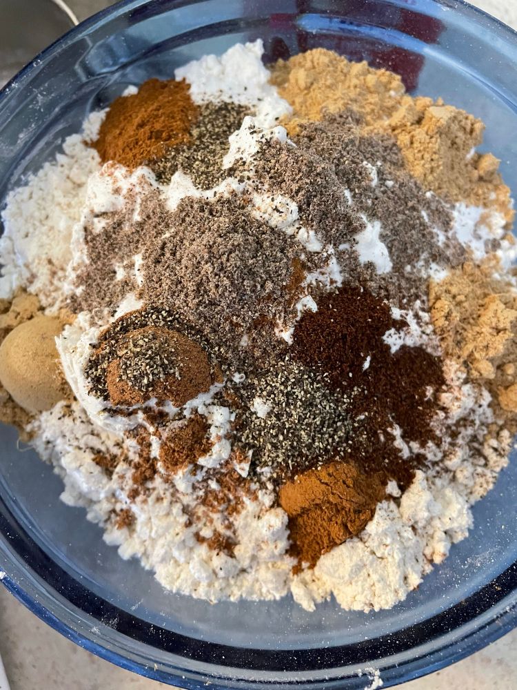 A blue glass mixing bowl filled with flour and ginger, cardamom, nutmeg, cinnamon, black pepper, and clove.