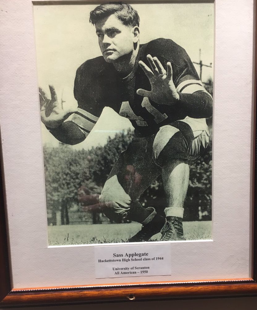A photo portrait of a Hackettstown High School class of 1944 football player named Sass Applegate, who was apparently a pretty big deal.
He's shown in a crouching position on a field backdropped with a grove of trees. His hands are outstretched, ready to catch a football in the town historical museum. Strong jawline and impressive hair parting game. The photo caption says, additionally, University of Scranton, All American
1950.