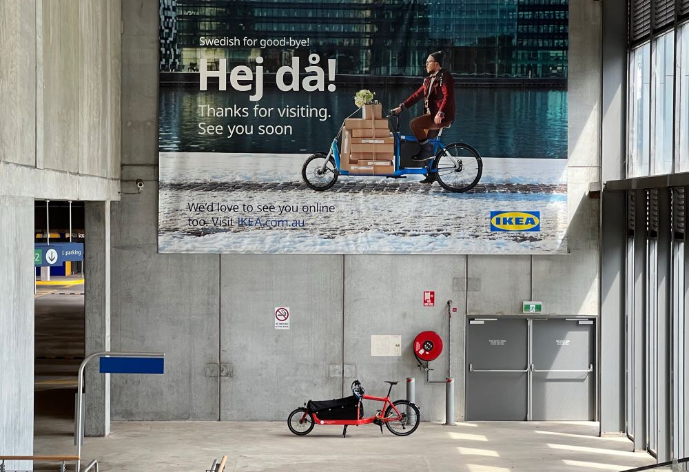 Bullitt cargo bike parked underneath an IKEA poster with a packed Bullitt cargo bike in an unused open area next to the entrance of the IKEA store in Melbourne.