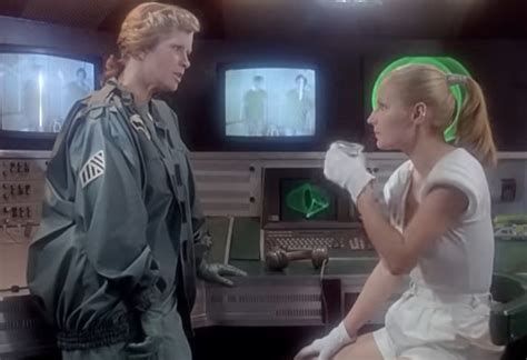 A scene from Seksmisja (1983). Lamia, a blonde woman dressed in white shorts, short-sleeved top and gloves, is sitting in a chair with a glass in her hand. Her superior, another blonde woman dressed in a green uniform, is standing in front of her, telling her something with a serious face. In the background, two men in green shirts are visible on a security screens placed above nondescript computer machinery.