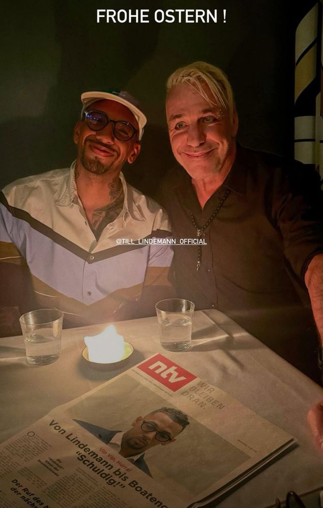 Bild der unheiligen Allianz von Jerome Boateng und Til Lindemann, wie sie vor einer Kerze und einer ntv-Zeitung mit einem Artikel über sich selbst sitzen. Jerome Boateng grinst dabei debil in die Kamera, während Lindemann mit grotesk verzerrtem Grinsen irgendwo ins Jenseits stiert.