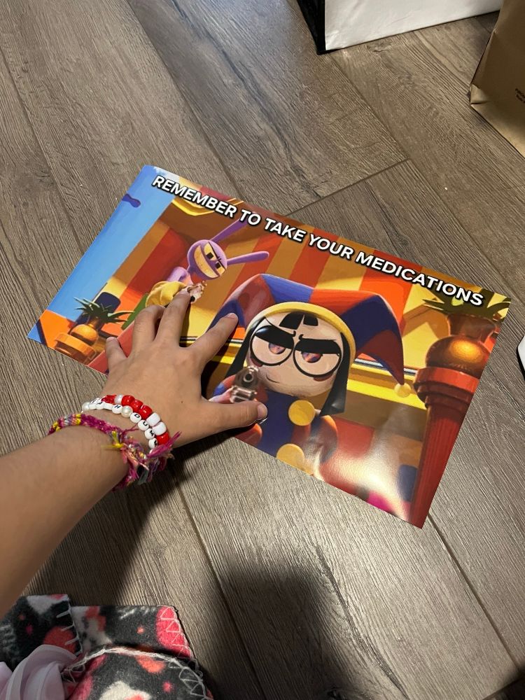 A hand clawing at a printed photo on the floor. The photo is a screenshot from The Amazing Digital with Jax, an anthropomorphic purple rabbit, and Pomni, a multicolored jester, each aiming a gun at the viewer. On the top of the photo there is blurry Impact font text that reads "Remember to take your medications"