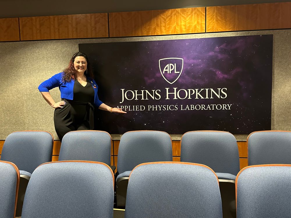 I am standing in front of the Johns Hopkins Applied Physics Laboratory logo banner with a purple space background. 