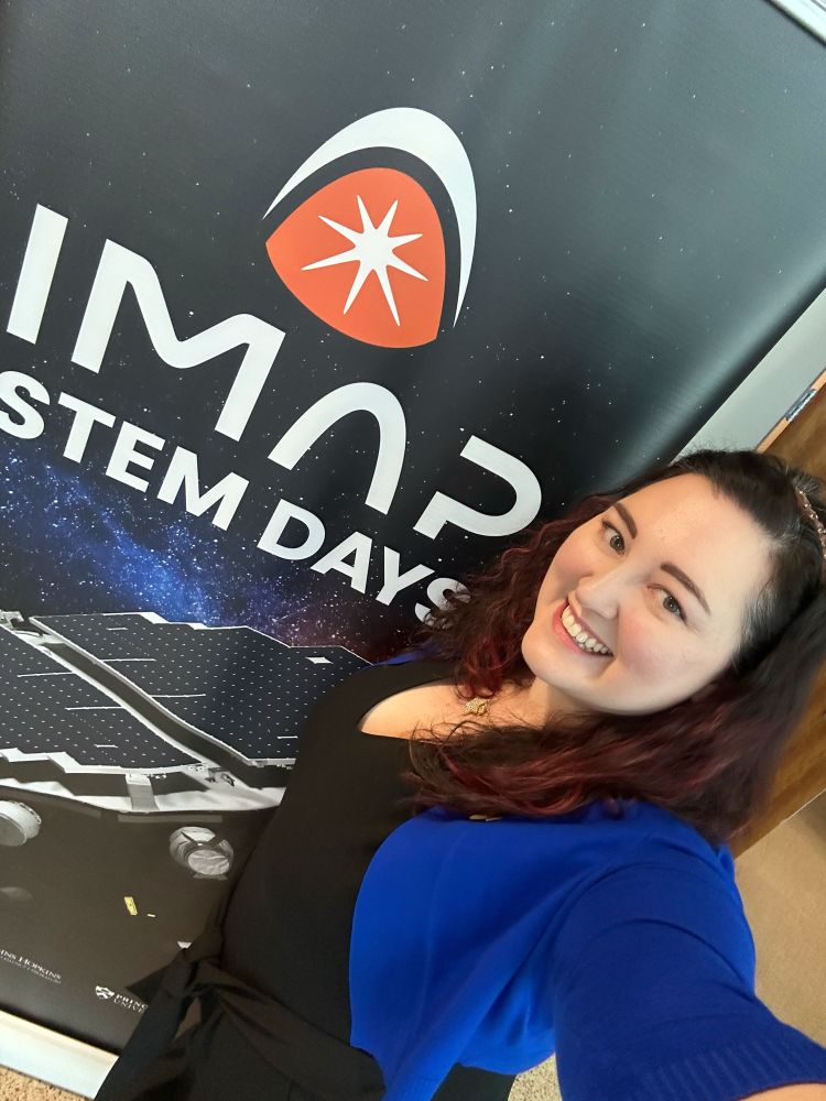 I am standing in front of a tall poster that says IMAP STEM DAYS with the orange and white IMAP logo and a digital rendering of the spacecraft below it. 
