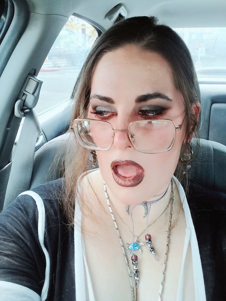 a selfie of me feigning a surprised expression while sitting in my car with gray seats. my makeup is silver/gray with bright red liner along my lower lids and dark brownish purple lipstick. my mouth is open and I'm looking down and to the left side of the frame so you can see the silver sparkles on my upper lid. im wearing a low bun, big gold glasses, several colorful necklaces, and a v neck dark gray hockey shirt with a grayish blue  under shirt.