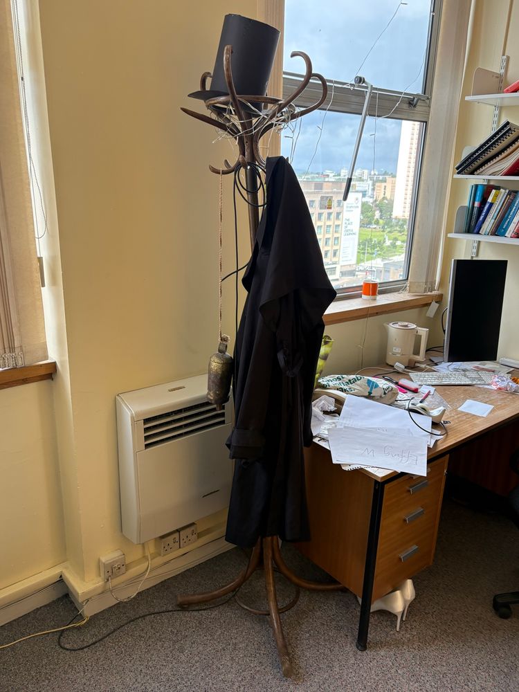 a wooden hatstand with a coat hanging on one of its hooks and a cardboard stovepipe hat on top, next to an untidy desk under an office window, with a pair of white stilettos tucked underneath the desk drawers