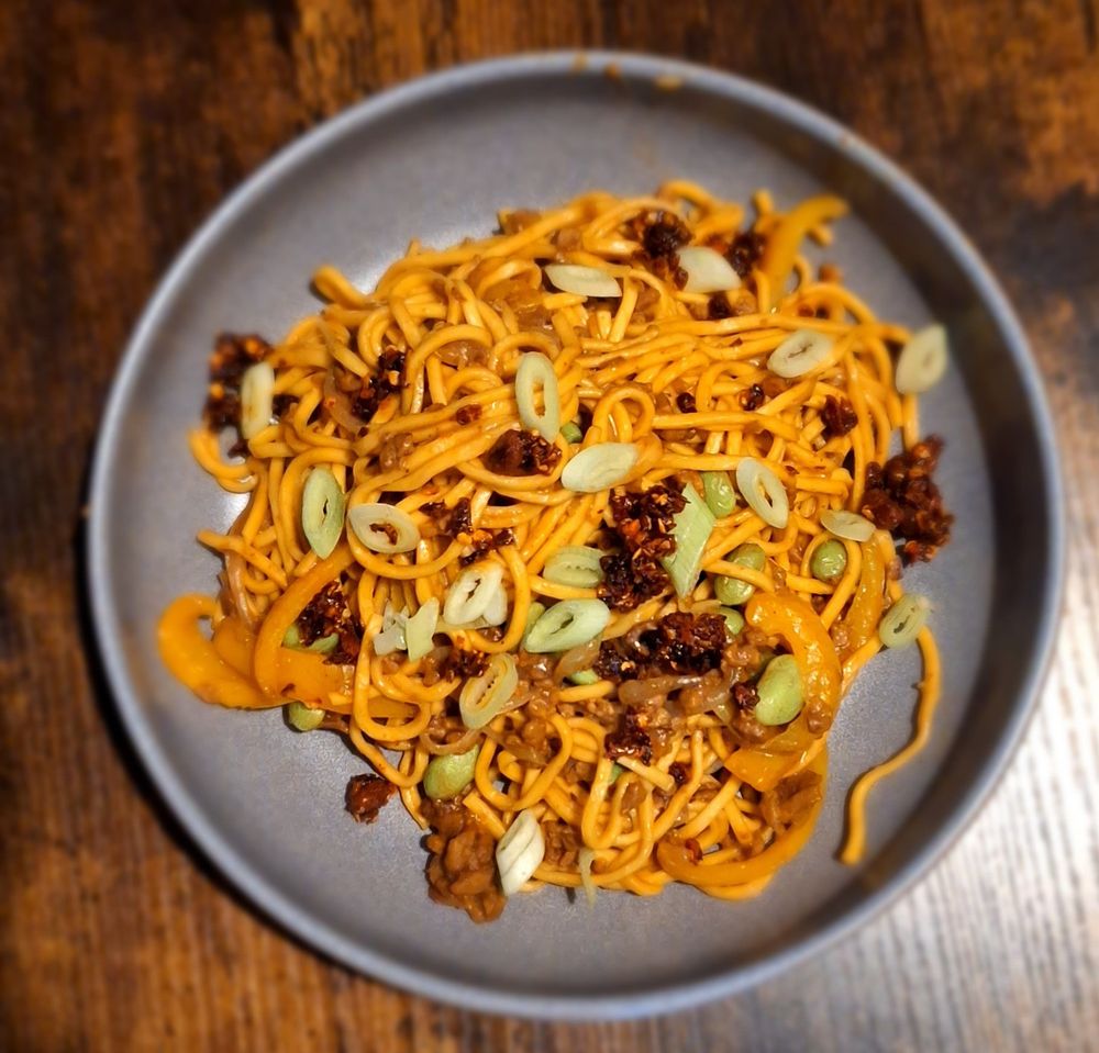 A grey bowl filled with yellow noodles, some soy mince, chilli crisp, spring onions. The bowl is sat on a dark brown table 