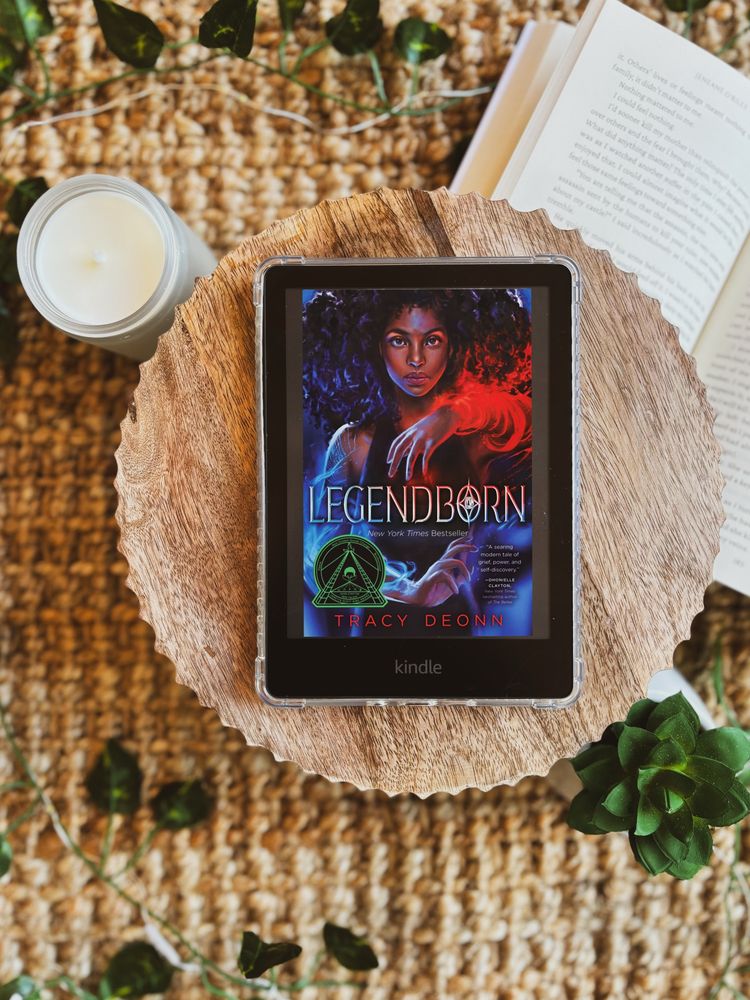 Legendborn book cover on kindle device, sitting on a wooden centerpiece, and surrounded by fake foliage, candle, and an open book.