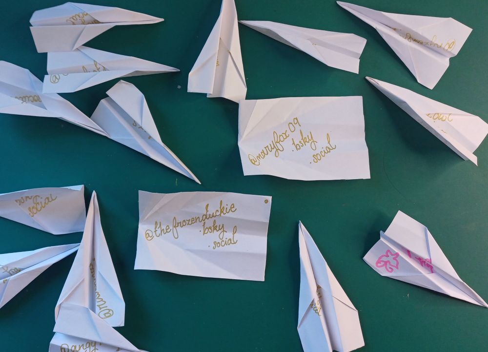 Image shows two creased pieces of paper with bsky handles written on them. One says @thefrozenduckie .bsky .social and one says @maryfox09 .bsky .social

They are surrounded by paper planes all with bsky handles on them. These are the winners of SunnyPeaceful's 100 follower art raffle.