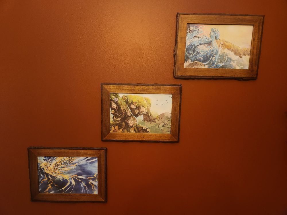 Three pieces of art (by artist Nolan Nasser, from the Spirit Island board game) in natural wood frames hanging on a red wall in a diagonal line, going from upper right to lower left. 