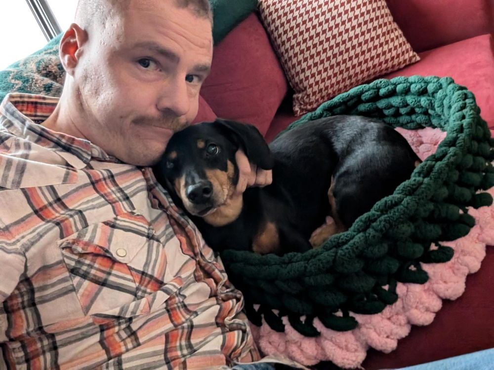 A selfie of my friend Commander Smith. He's looking at the camera to his right, while cuddling his doggos Miss Morita in her dog bed. He's wearing a light coloured button shirt. The dog bed is green and pink.