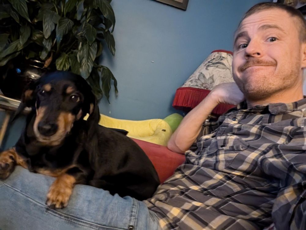 A photo of my friend Commander Smith He is smiling at the camera with one of his doggos on his lap. He's wearing a blue patterned button shirt and light blue jeans. The doggo is Miss Morita, she is a back and tan daschund.
