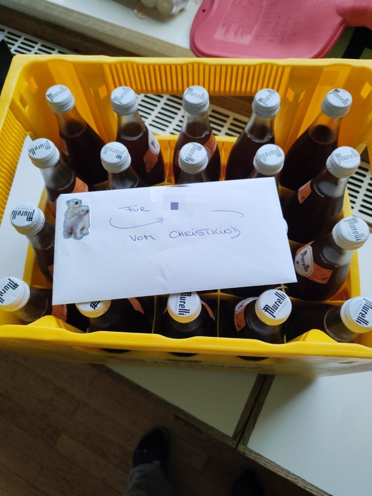 Ein gelber Kasten mit 20 Flaschen Spezi und einem weißen Kuvert darauf steht: 
Für (mein Name) 
Vom Christkind