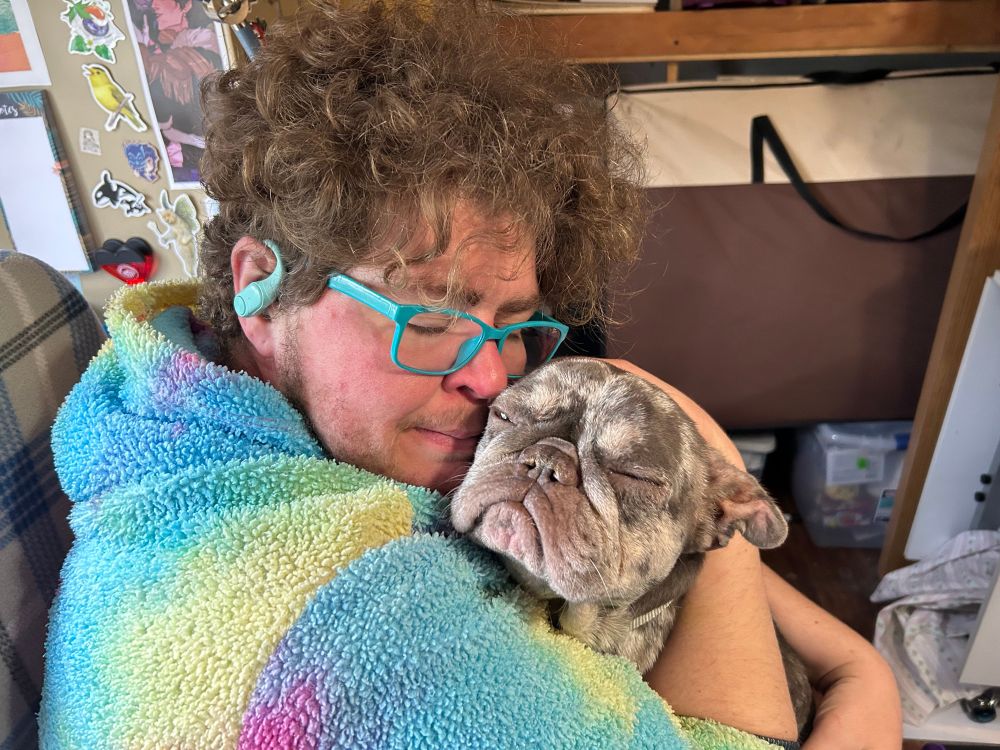 Brown, curly haired man with teal glasses and AirPods and a tyedyed hoodie hugs the squish faced dog. Her eyes are closed in contentment. The phantoms are at bay- for now. 