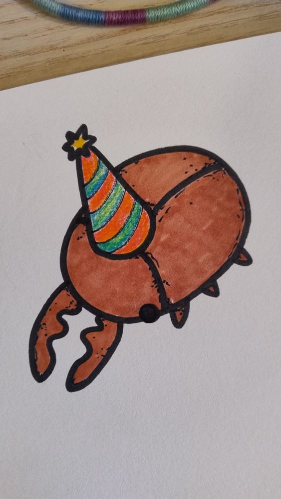 A cartoony drawing on grainy paper of a chibi stag beetle wearing a party hat. The outline of the bug is done in black sharpie, the beetle is facing left. He is light brown colored and the party hat has a yellow pompom and orange-green stripes. 