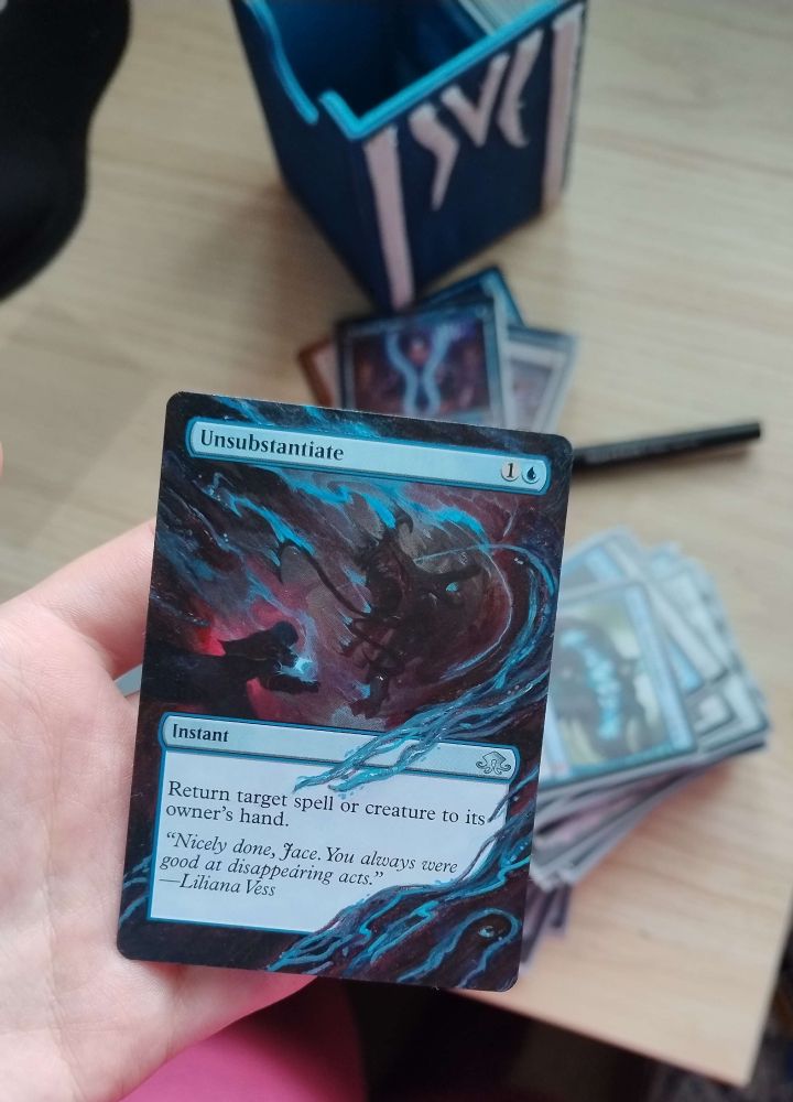 A picture of someone holding an altered MTG card: Unsubstantiate. The borders have been extended to also show the magical effect above the textbox and to the sides. In addition, magical effects have been added to partially cover the rules text box and the bottom right corner, replicating the blue magics already present in the original art. Out of focus, in the background, a stack of other Jace cards lying on the desk is visible. 