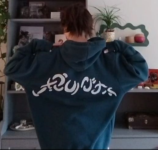 A photo of a person with their back to the camera. The person is wearing an oversized teal blue hoodie. There are mystical, swirly white symbols stitched onto the back of the hoodie in a slight arc. These are the magical symbols that are also visible between the hands of Jace in the art on the original printing of "Jace, The Mind Sculptor" card. 