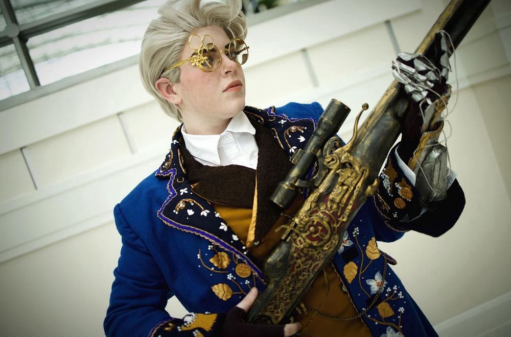 A light grey haired man with ear length hair and round golden spectacles complete with jewelers lenses. He wears a white button up shirt, and golden vest with a dark brown contrast collar, and a royal blue coat lavishly decorated in golden leaves, embroidery, and white organza flowers. The coat’s collar and sleeve cuffs are a contrast of navy blue and smaller embroidery designs. He holds up a long brown rifle that contains red and gold designs and a large vision sight. His left hand is covered by a silver and gold metallic glove. 