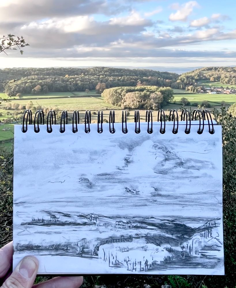 This is a photograph from the top of the hillside looking down into a valley. The Sun is low the sky causing long cast shadows and in the foreground is a drawing of the same scene.