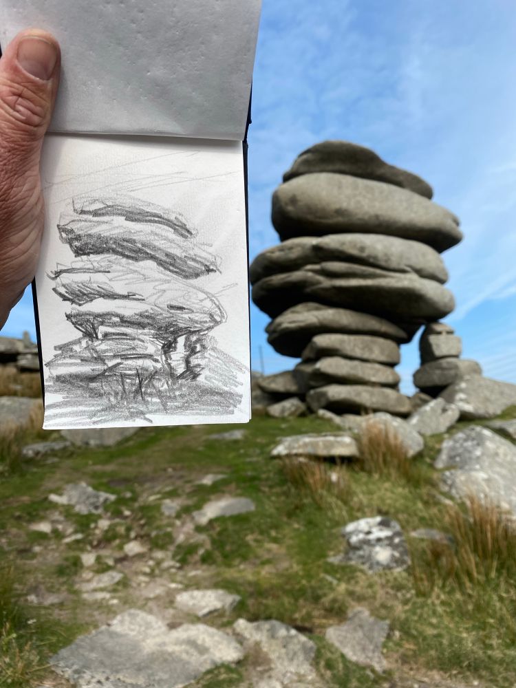This is a photograph of a naturally occurring stack of boulders. In the foreground is someone holding up the sketchbook with the same scene drawn on it.. this was taken in Cornwall at the cheesewring. 