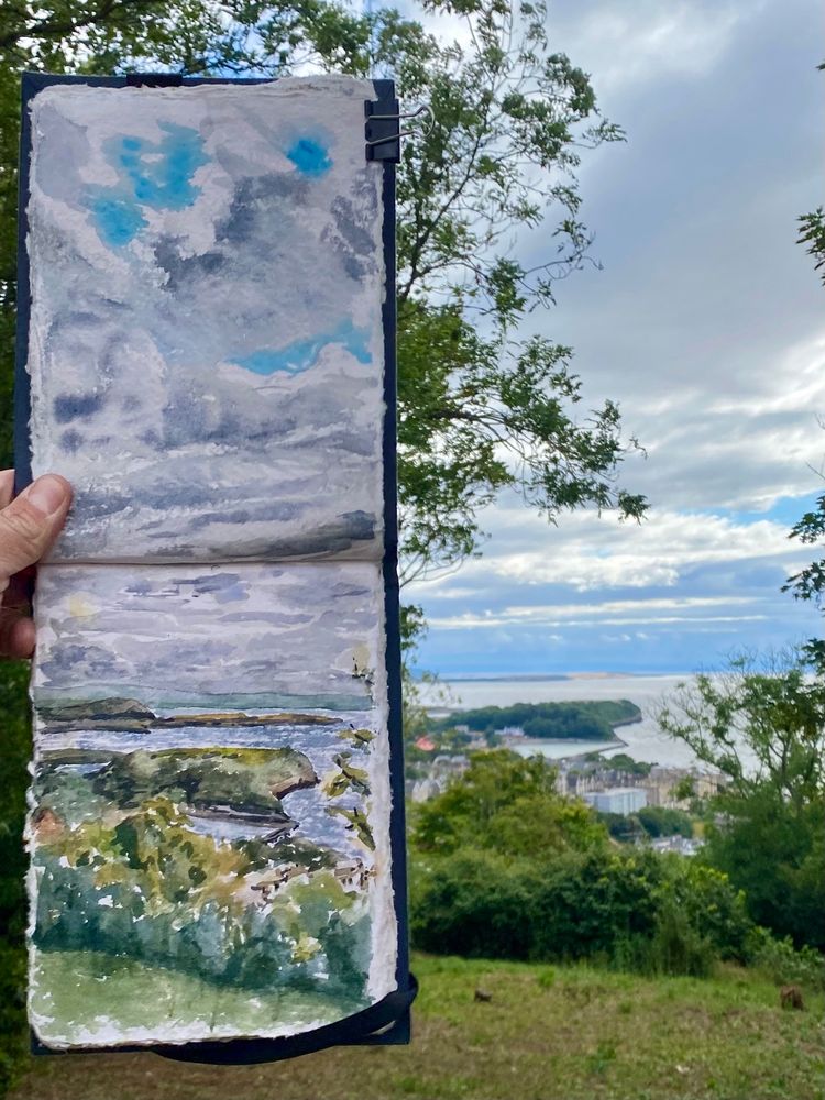 This is a photograph of a coastal scene from the point of view on top of the hillside. In the photo you can also see a sketchbook that's painted the same scene. The painting is a watercolour that has been done loosely. The majority of the painting is the sky.