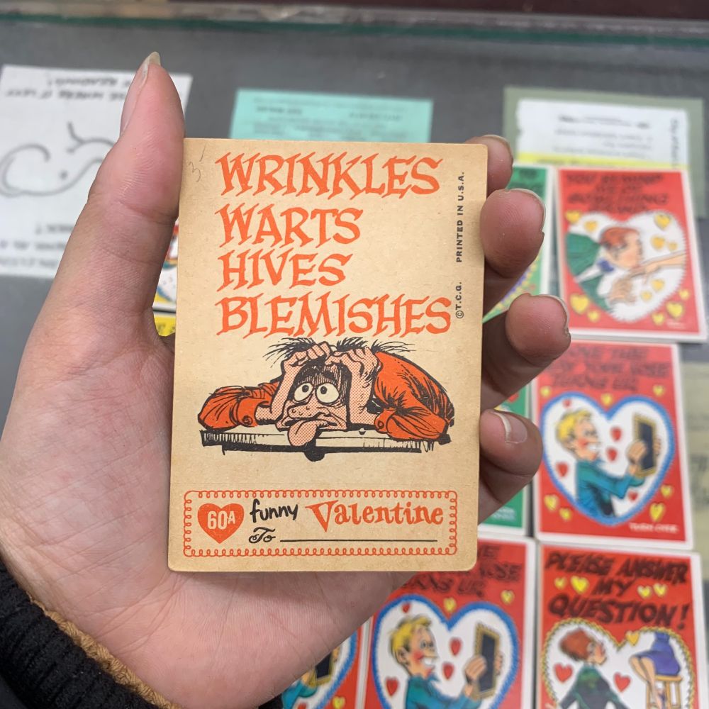 The back side of a vintage Valentine, which reads "Wrinkles, Warts, Hives, Blemishes" with the cartoon of a person holding their head below it. This is the back side of the vintage Valentine from the photo before it.