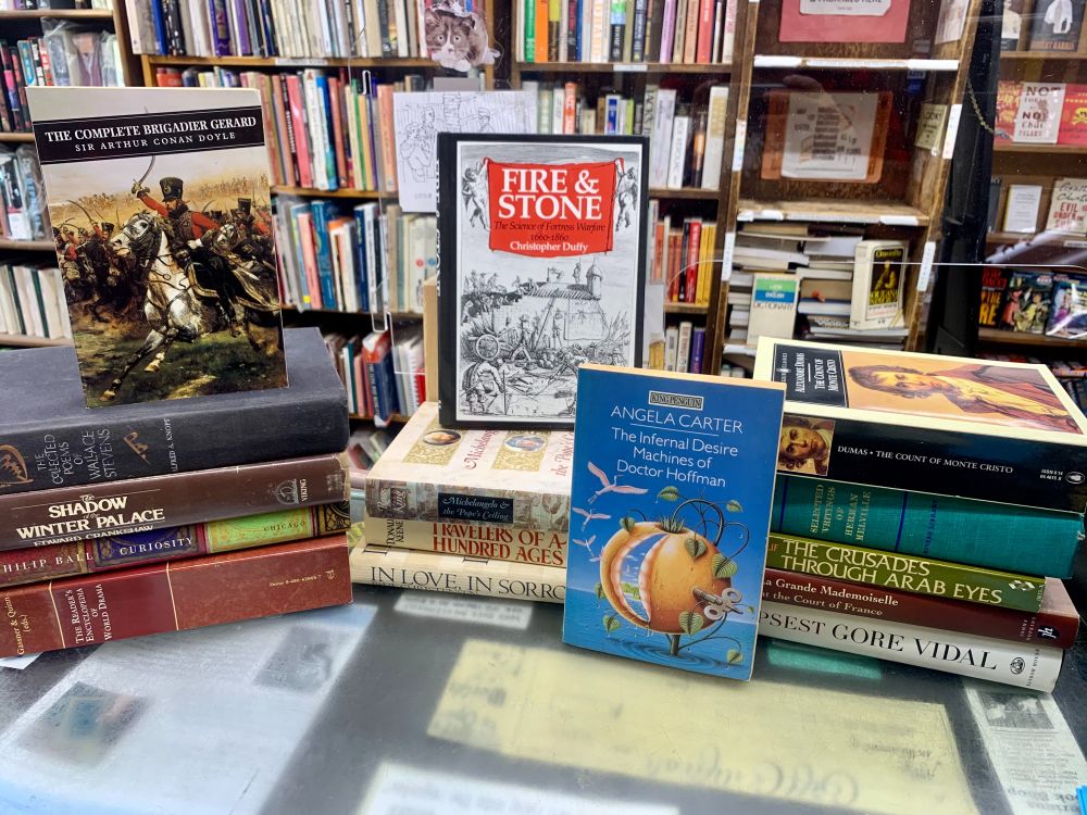 Three stacks of books on the counter at Curious Book Shop. The face out titles include Fire and Stone by Christopher Duffy and The Infernal Desire Machines of Doctor Hoffman by Angela Carter.