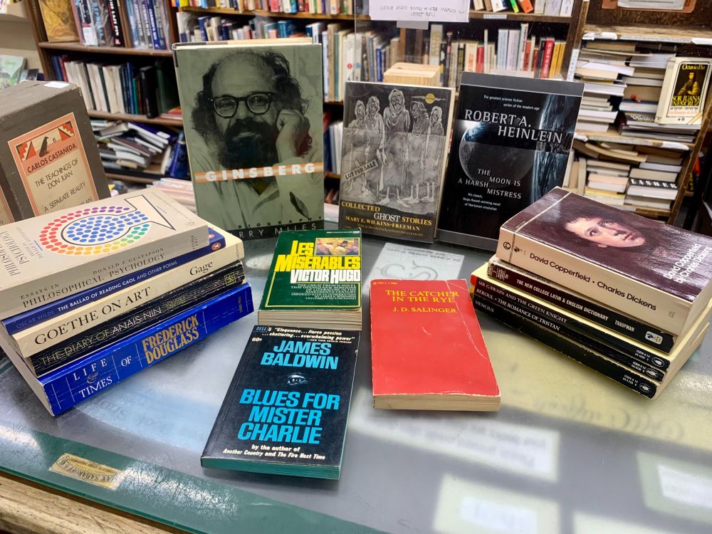 A stack of used books on the counter at Curious Book Shop. Some of the included titles are Blues for Mister Charlie by James Baldwin, The Catcher in the Rye by J.D. Salinger, The Moon Is A Harsh Mistress by Robert Heinlein, and Goethe on Art.