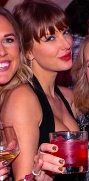 A somewhat close-up picture of a blonde woman with bangs (Taylor swift) and two women on either side of her who have been cropped out. Taylor is wearing a black halter dress with her hair in a ponytail and a mischievous look on her face. She is holding a red/pink colored iced drink that might be a cocktail up to the camera. This picture is widely assumed to be from her 35th birthday party. 
