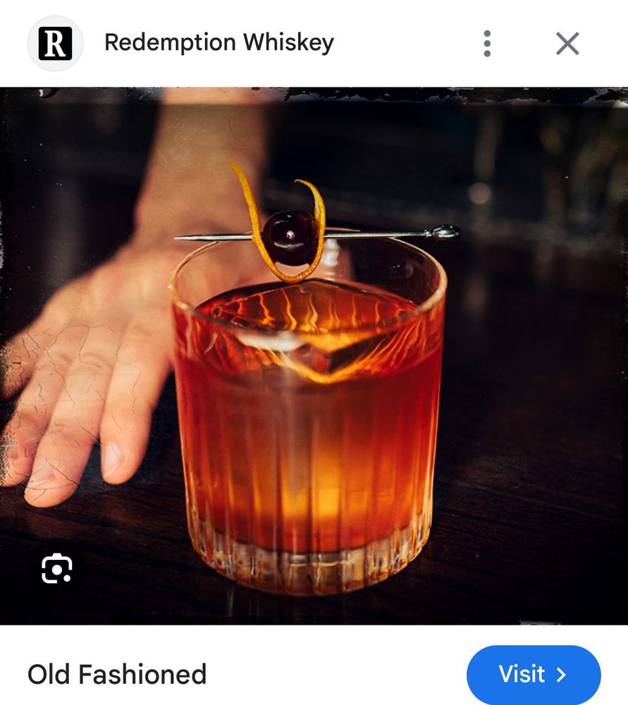 A screenshot of a commercial picture of an old fashioned cocktail. The cocktail is the main focus and is a warm-toned copper color with a single large cube of ice and a garnish of cherry and orange peel on a drink skewer, laid across the top of the glass. A person’s hand is also shown laying on the table beside the glass. The table and background are hard to see but are dark in color. 