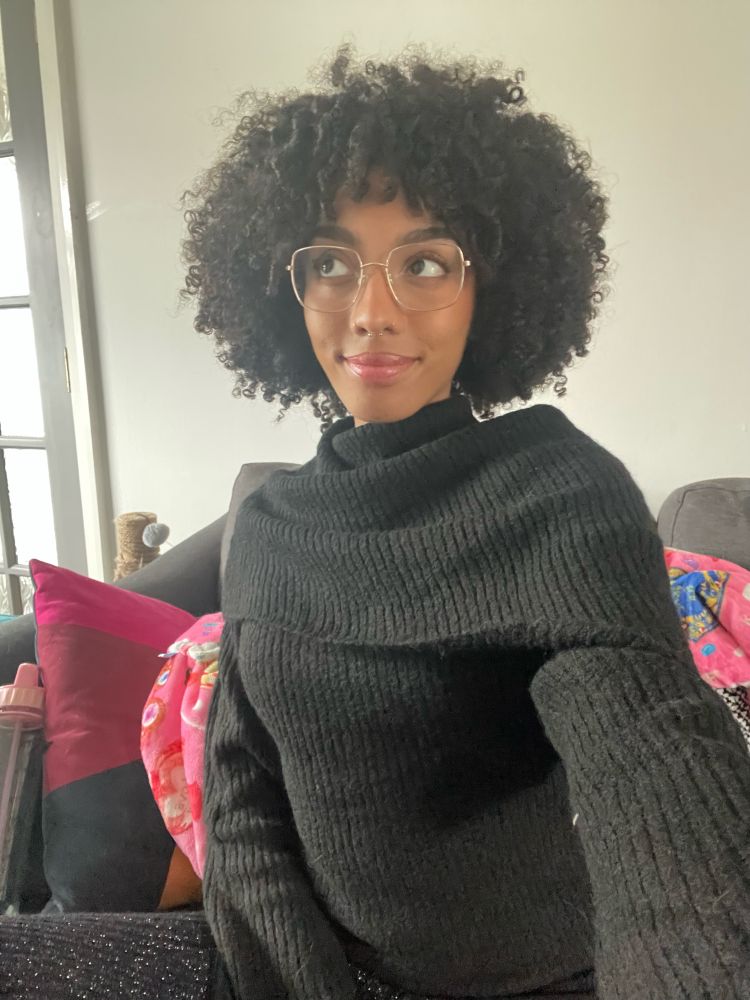 Selfie of Chora, a black woman with afro curly hair and gold glasses in a black sweater, glittery leggings sat on her sofa.