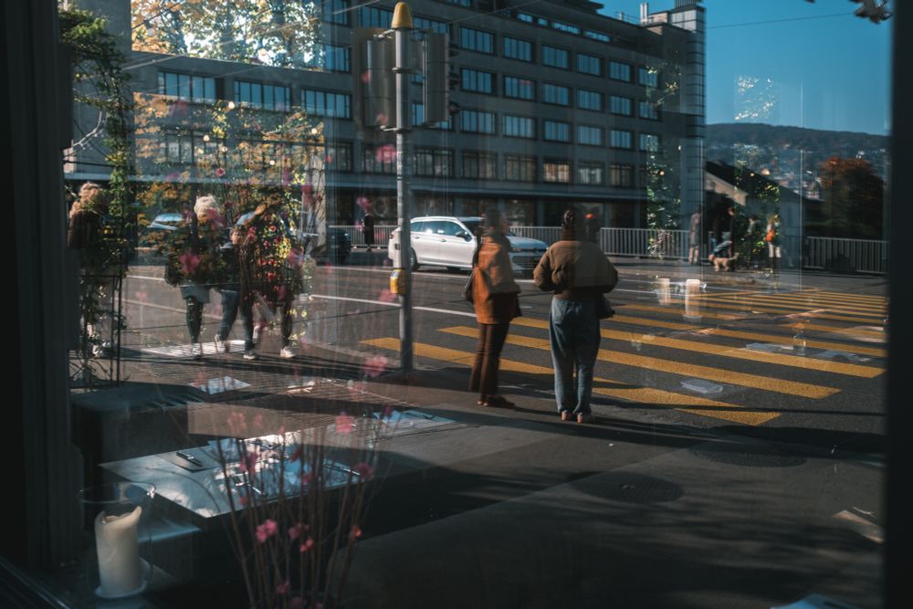 „Straßenszene durch ein Schaufenster: Herbstbäume, Passanten und ein vorbeifahrendes Auto verschmelzen mit der Spiegelung von Blumen und Kerzen – Innen und Außen in poetischer Balance.“