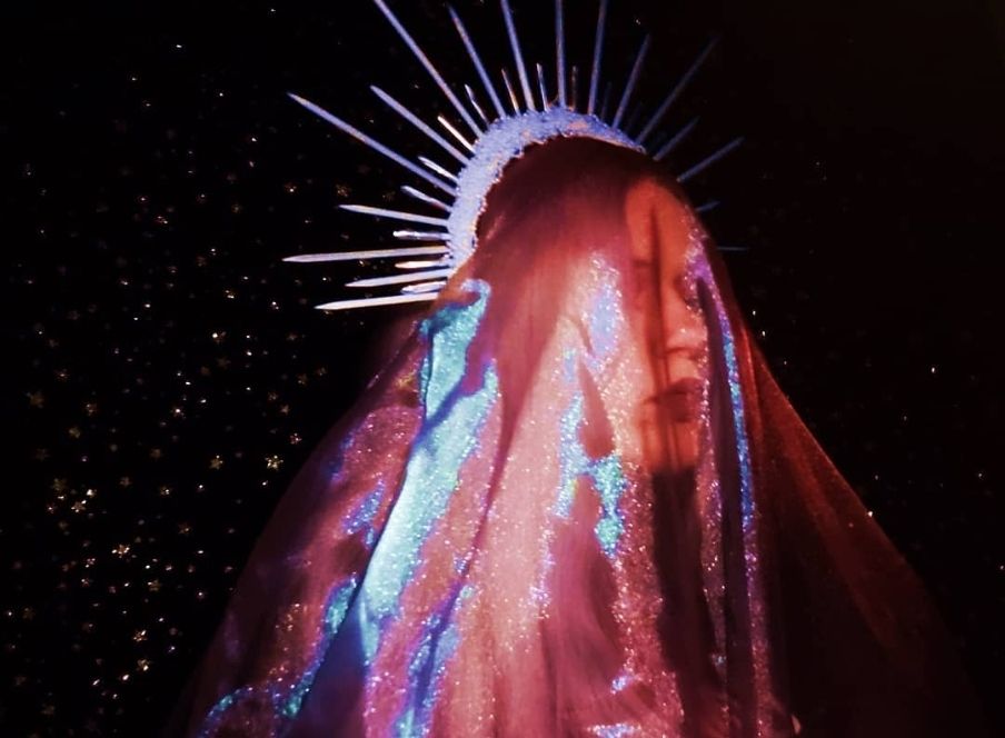 Woman in a red/blue veil with a spike-y crown in front of a background of stars.