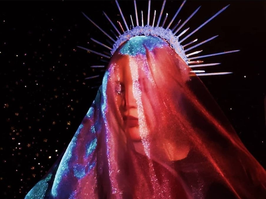 Woman in a red/blue veil with a spike-y crown in front of a background of stars.